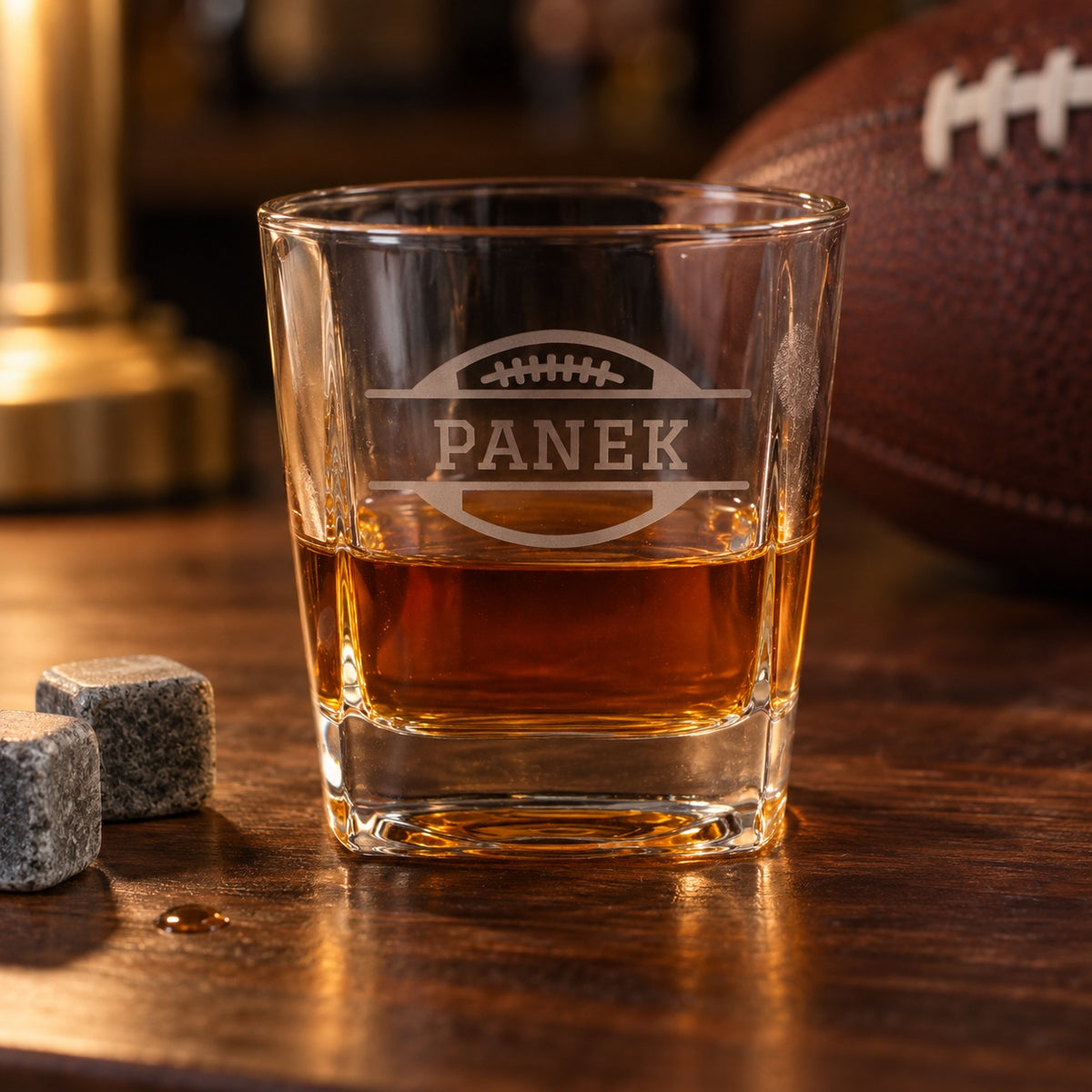 Bourbon Blitz personalized football whiskey glass etched with PANEK across a football, a square bourbon glass with whiskey stones on a bar