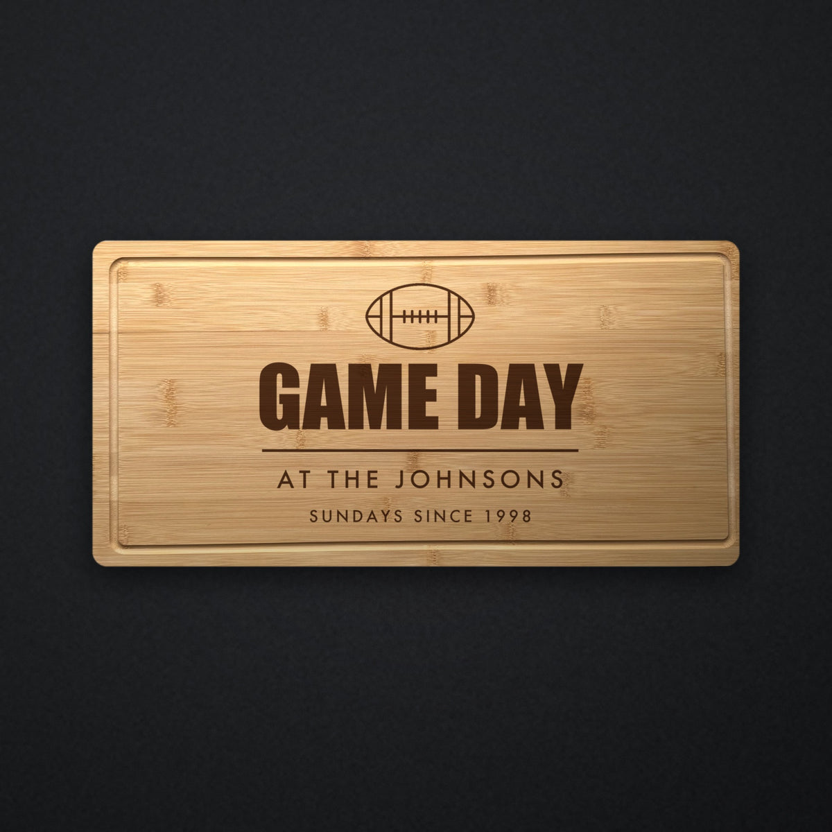 Personalized game day cutting board in solid bamboo on dark slate, engraved with a football above Game Day, the family name and the year.