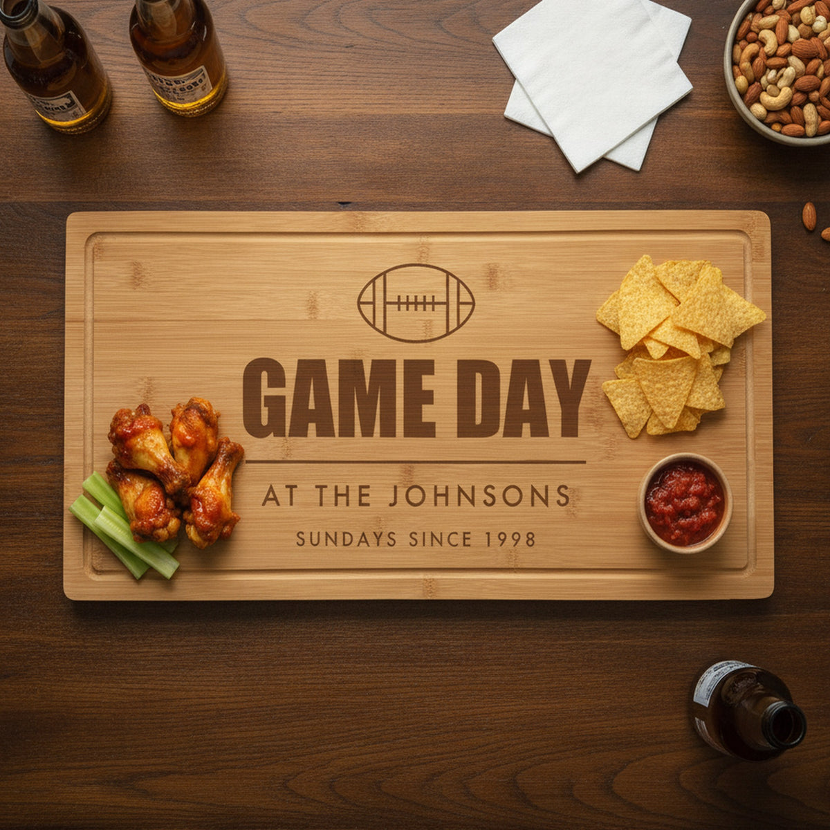 Engraved Game Day bamboo board on a wood table with buffalo wings and celery at one end, tortilla chips and salsa at the other, and two beers.