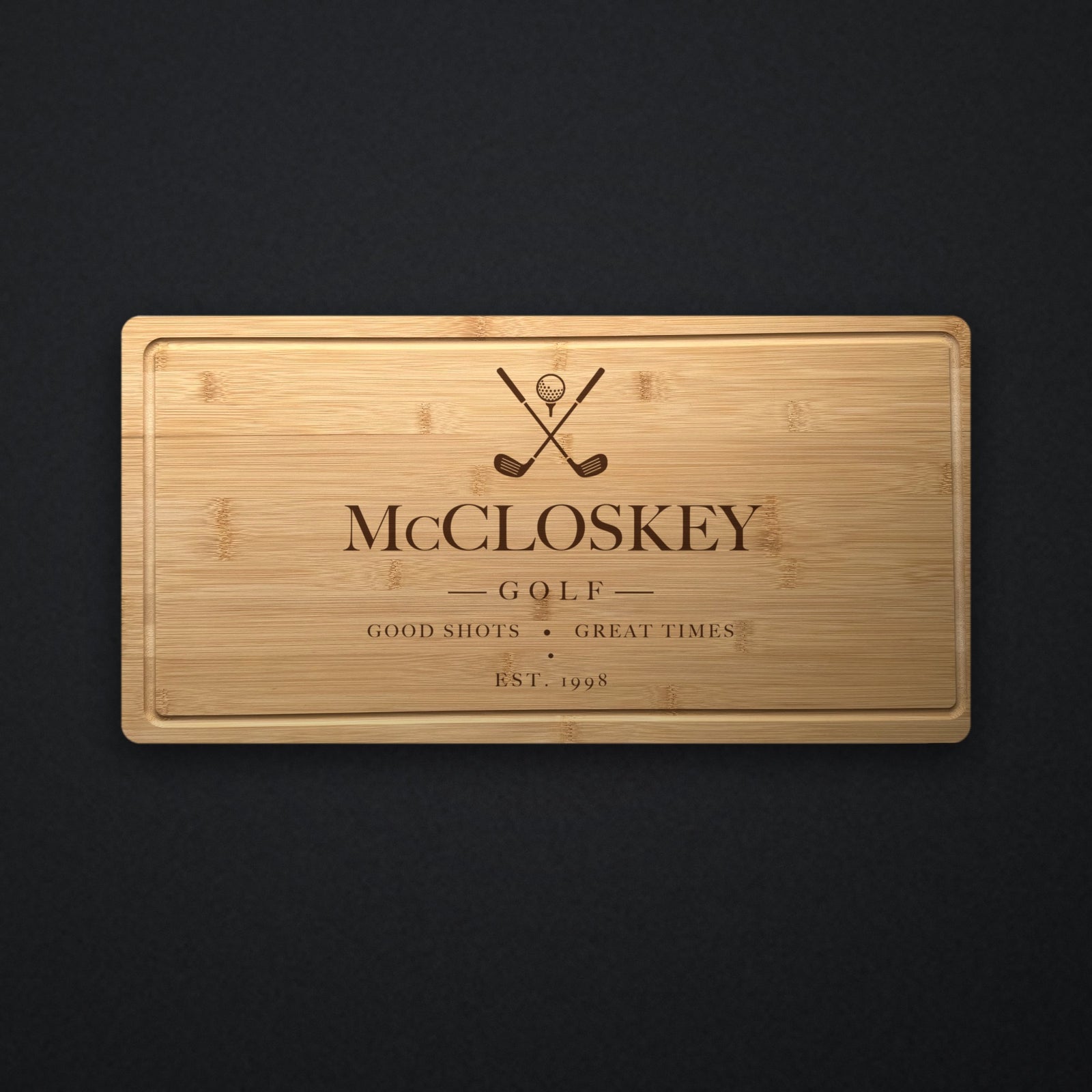Personalized golf cutting board in solid bamboo on dark slate, engraved with crossed golf clubs and a ball above the family name and the year.