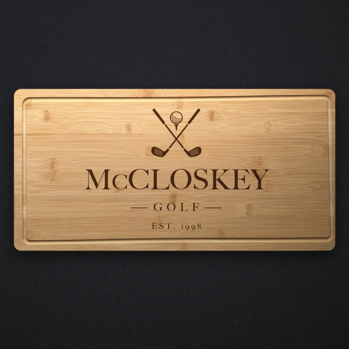 Personalized golf cutting board in solid bamboo on dark slate, engraved with crossed golf clubs and a ball above the family name and the year.