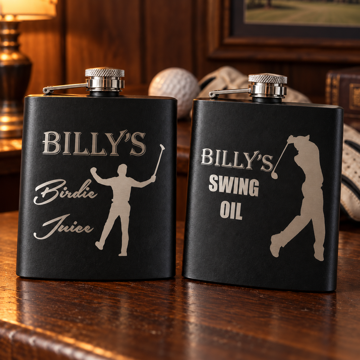 Two personalized golf flasks in matte black, one engraved Billy's Birdie Juice and one Billy's Swing Oil, on a dark walnut bar with a golf ball behind