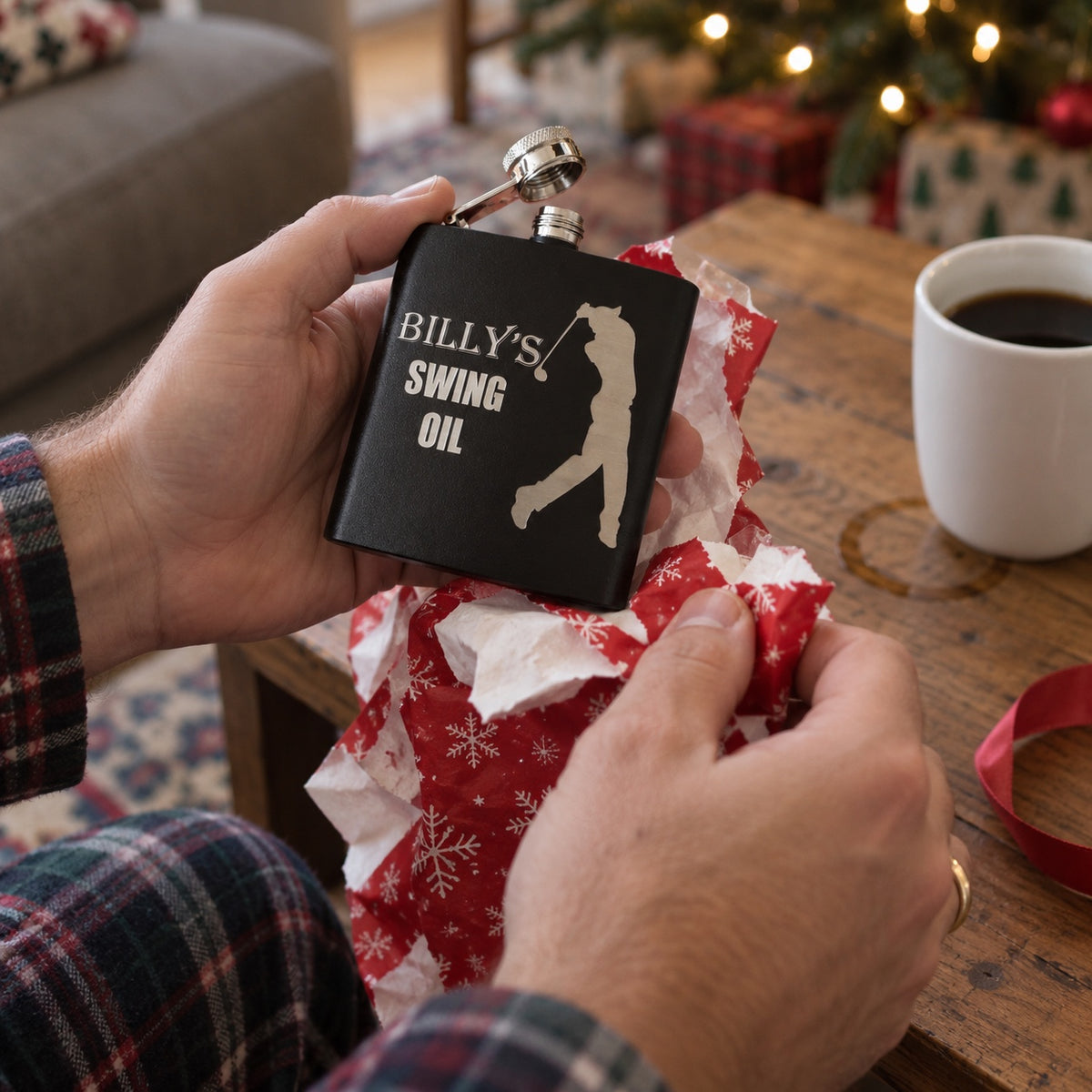 A man unwraps a personalized Swing Oil golf flask from red Christmas wrapping paper by the tree, a golf gift for him on Christmas morning