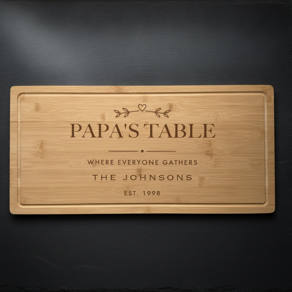 Personalized grandpa cutting board in solid bamboo on dark slate, engraved Papa's Table above the family name and the year, with a small sprig and heart.
