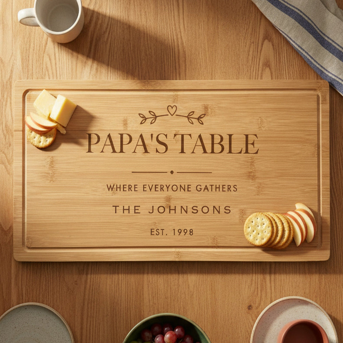 Engraved Papa's Table bamboo board on a warm oak family table with mugs, plates, grapes and cheese and crackers set at each end of the board.