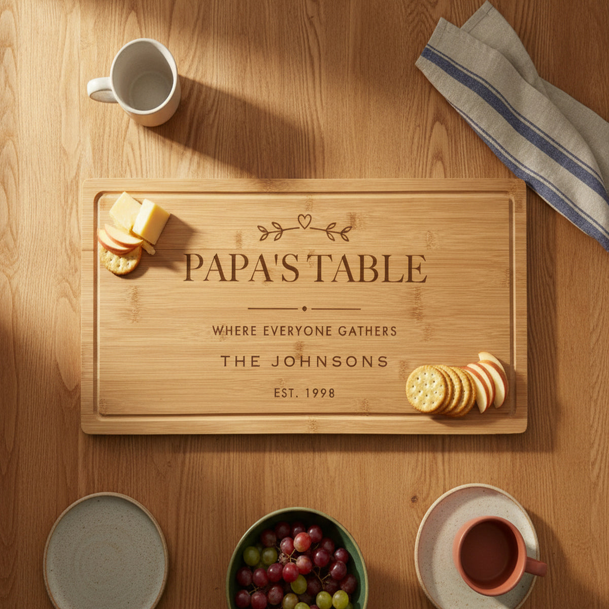Engraved Papa's Table bamboo board on a warm oak family table with mugs, plates, grapes and cheese and crackers set at each end of the board.