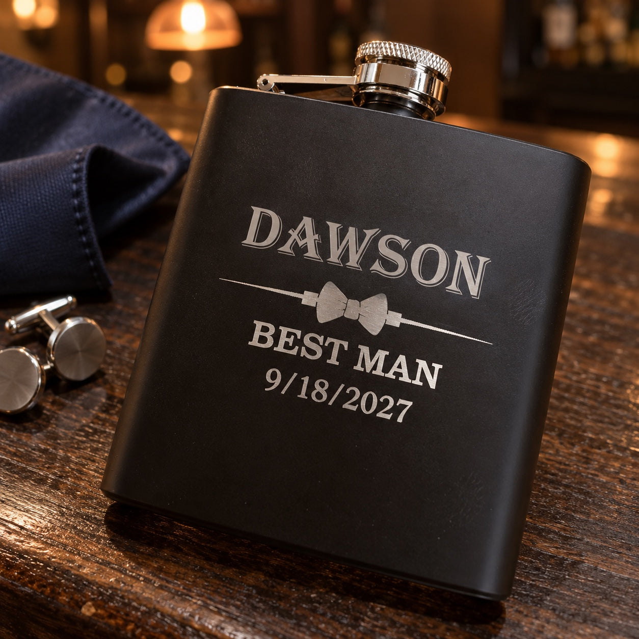 Personalized groomsmen flask in matte black engraved DAWSON above a bow tie, BEST MAN and 9/18/2027, lying on a walnut bar beside silver cufflinks.
