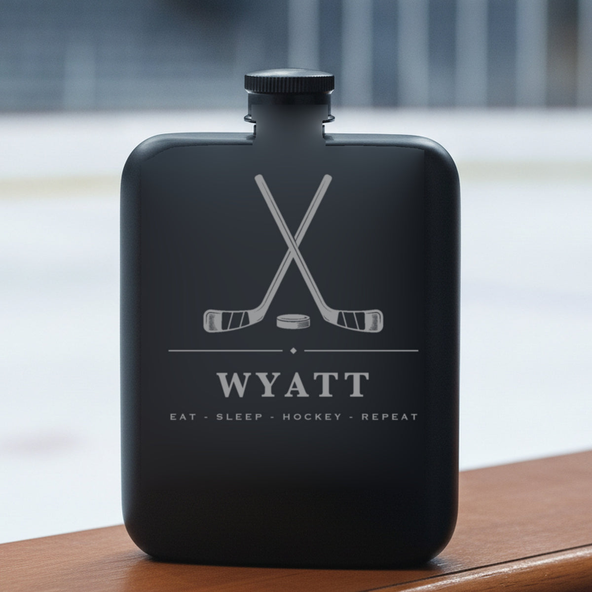 Personalized hockey flask engraved Eat - Sleep - Hockey - Repeat standing on a rink dasher board with the ice blurred behind.