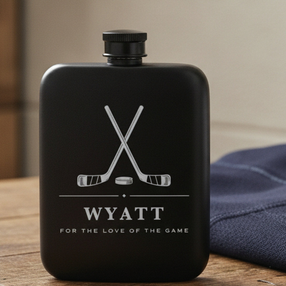 Engraved hockey flask reading For The Love Of The Game on a locker room bench beside a folded navy team jersey.