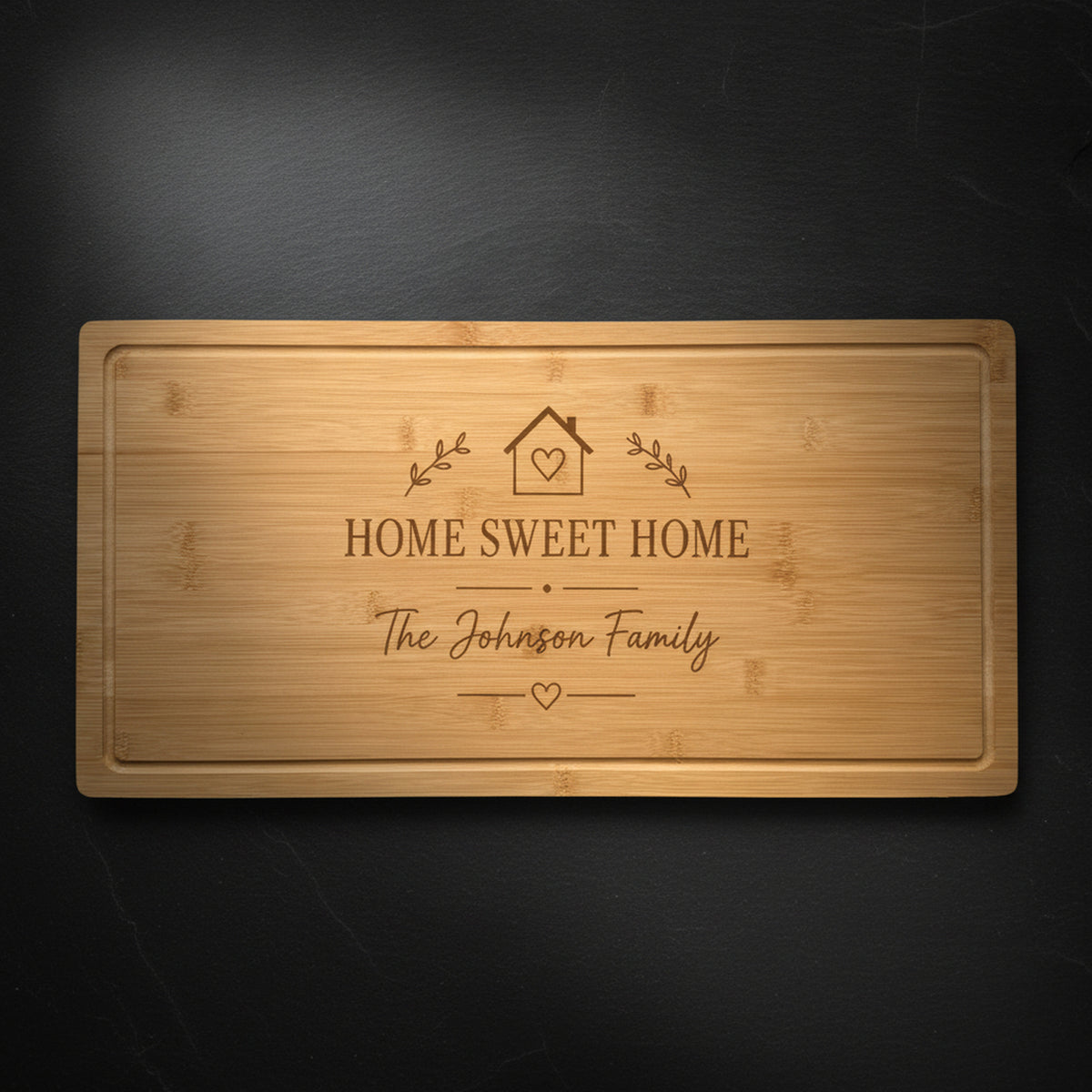 Personalized housewarming cutting board in solid bamboo on dark slate, laser engraved with a Home Sweet Home crest above the family name.