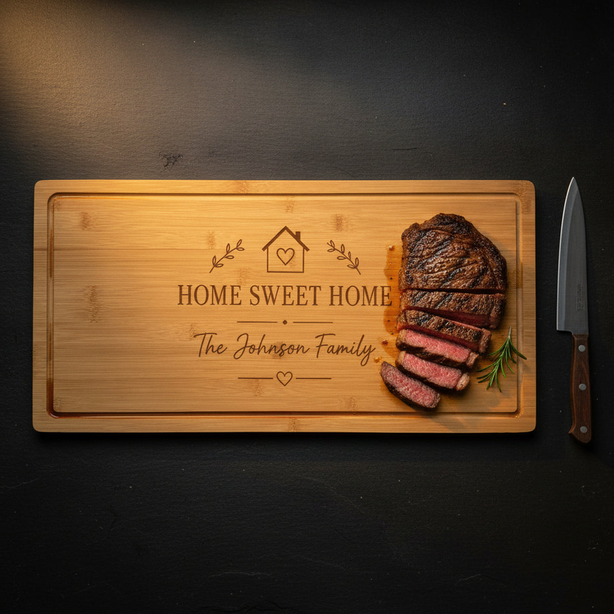 Large engraved bamboo carving board on dark slate holding a sliced medium rare ribeye with rosemary, juices caught by the carved drip ring, chef knife alongside.