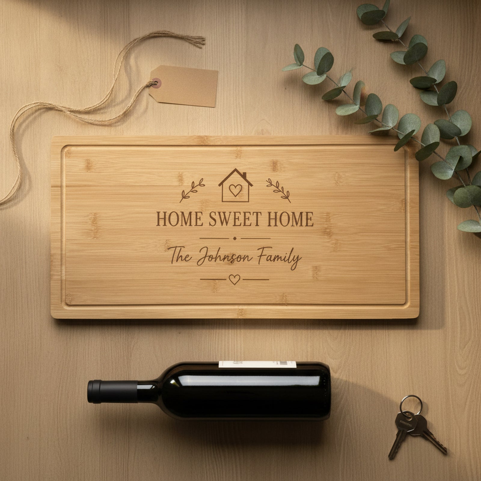 Personalized new home cutting board on an oak counter with jute twine, a kraft gift tag, a bottle of wine, eucalyptus and a set of house keys.