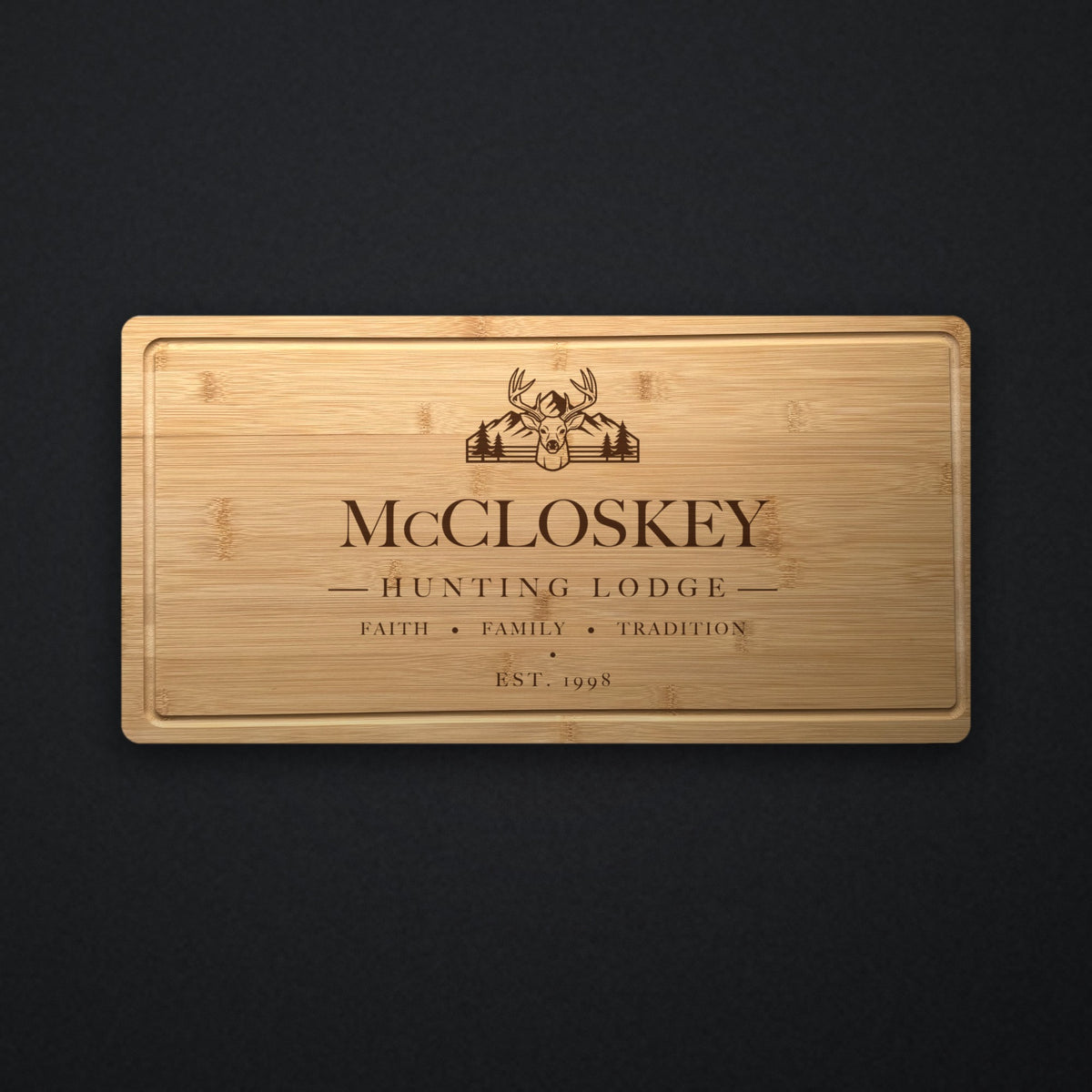 Personalized hunting lodge cutting board in solid bamboo on dark slate, engraved with a buck, mountains and pines above the family name and the year.