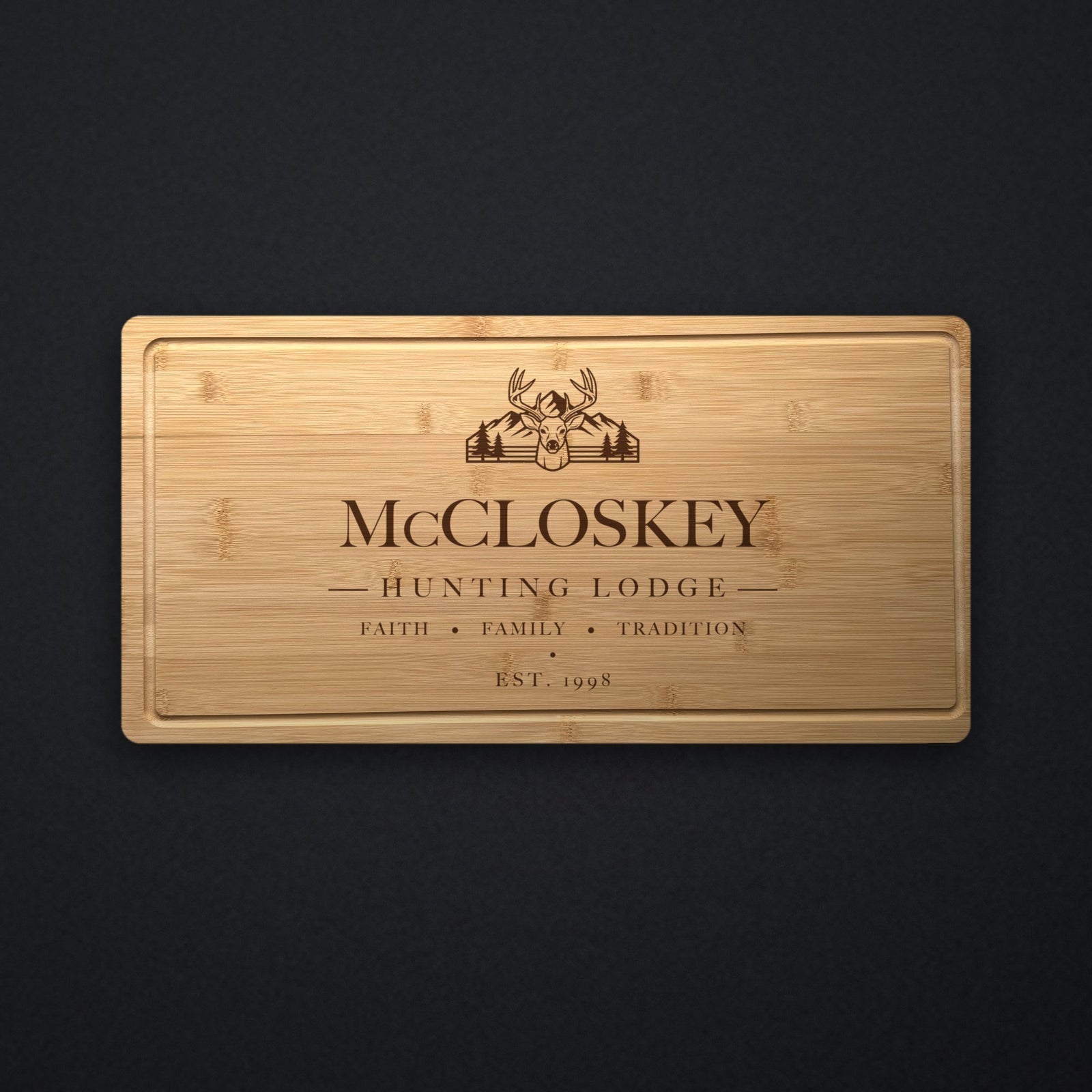 Personalized hunting lodge cutting board in solid bamboo on dark slate, engraved with a buck, mountains and pines above the family name and the year.