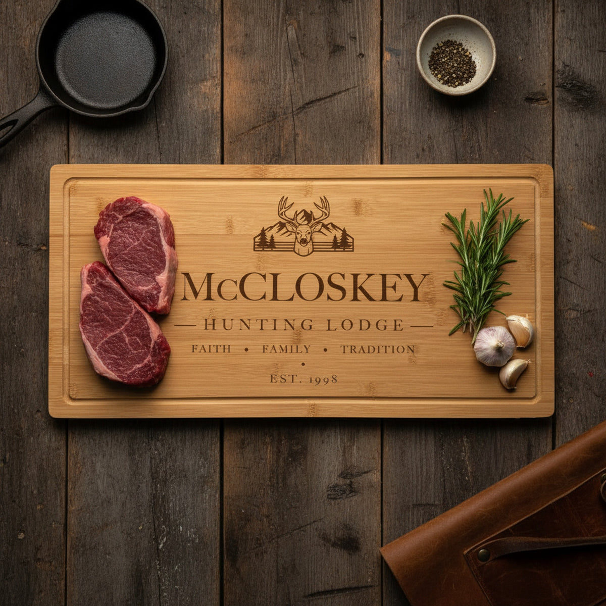 Engraved hunting lodge bamboo board on dark barn wood with two thick venison steaks at one end, rosemary and garlic at the other, beside a cast iron skillet.