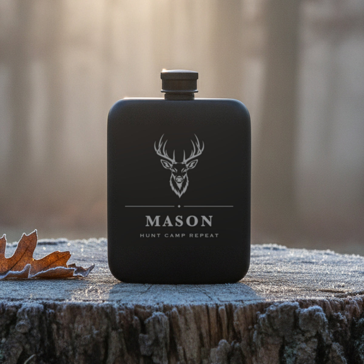 Personalized buck engraved hunting flask reading Hunt Camp Repeat, standing on a frosted oak stump at first light in misty woods.