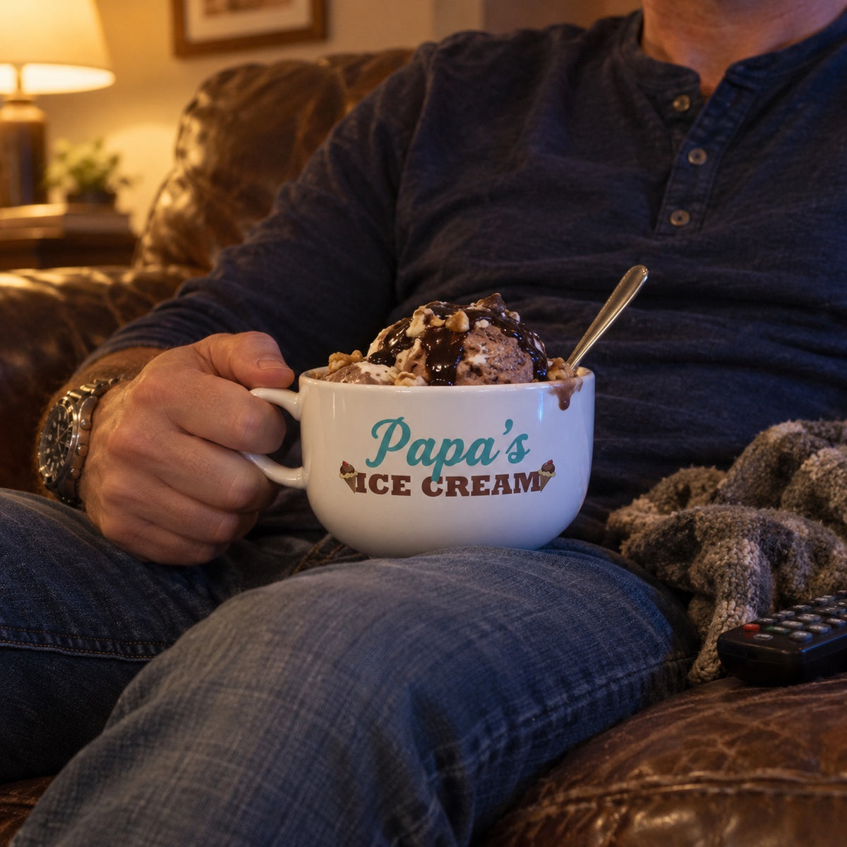 A man relaxes on a leather couch holding a personalized ice cream bowl printed with Papa's, filled with a hot fudge and walnut sundae