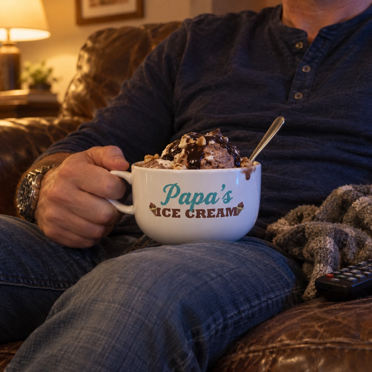 A man relaxes on a leather couch holding a personalized ice cream bowl printed with Papa's, filled with a hot fudge and walnut sundae