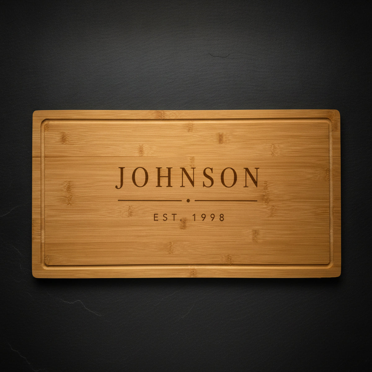 Personalized last name cutting board in solid bamboo on dark slate, laser engraved JOHNSON above EST 1998 in clean block type.
