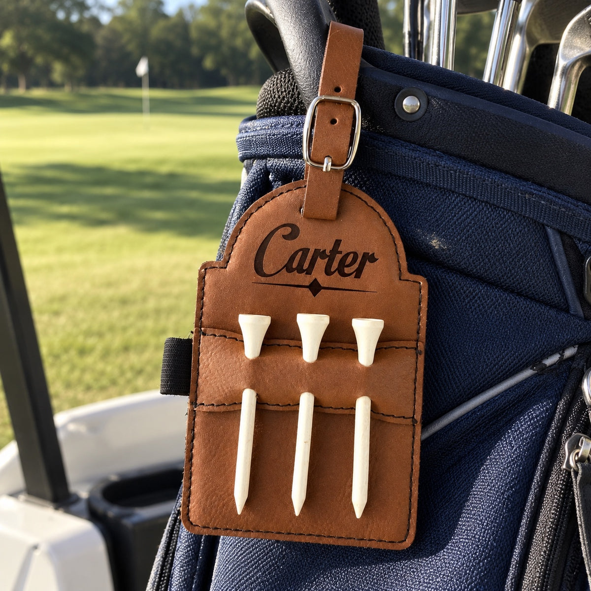 Personalized leather golf bag tag engraved Carter with three white tees, buckled onto a navy golf bag on a cart by the fairway