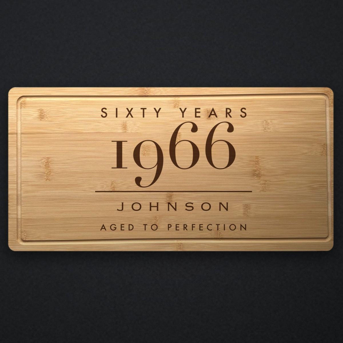 Personalized milestone birthday cutting board in solid bamboo on dark slate, engraved Sixty Years above the birth year, the name and Aged To Perfection.