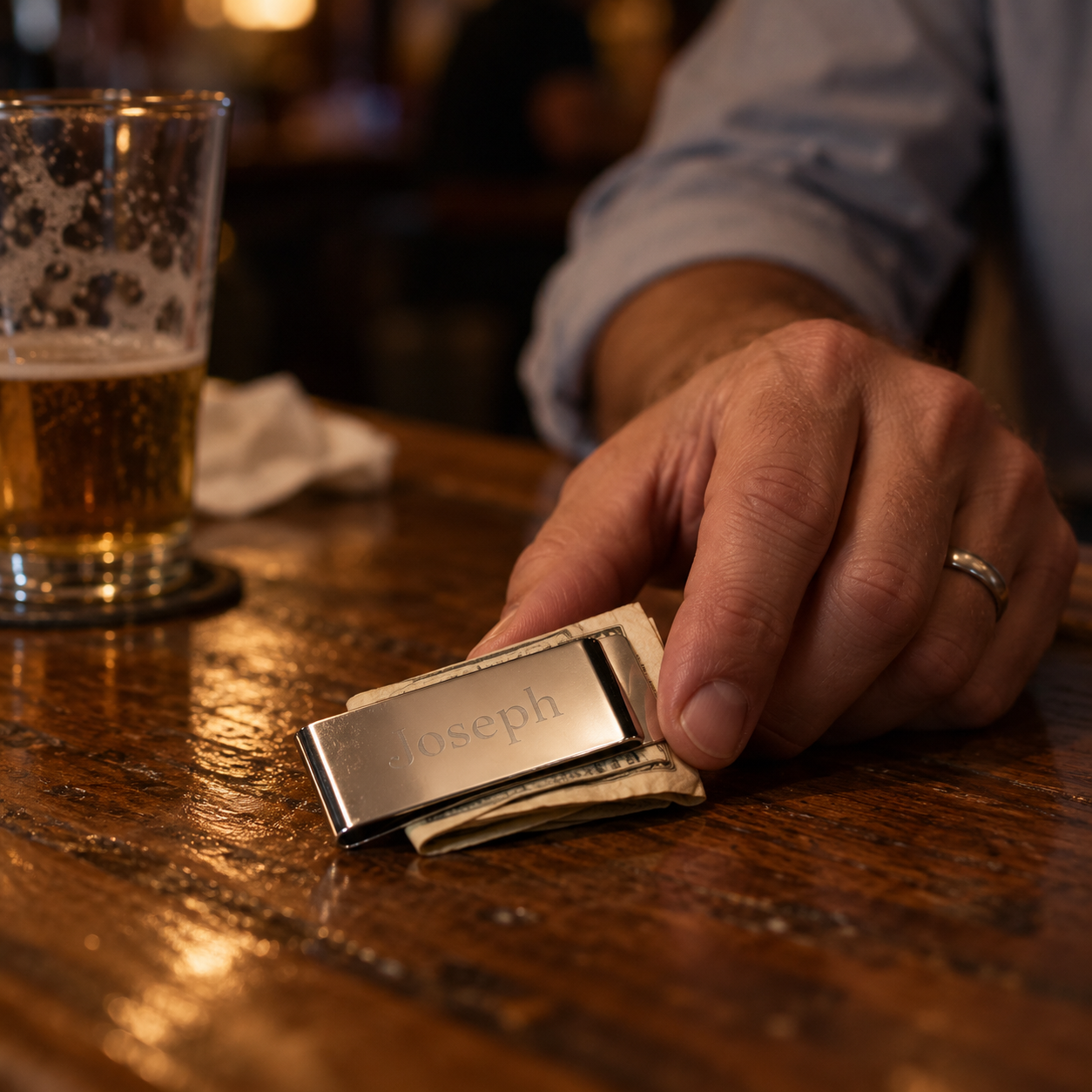 A man sets his personalized money clip engraved Joseph on the bar to pay the tab, folded cash held in the mirror steel clip