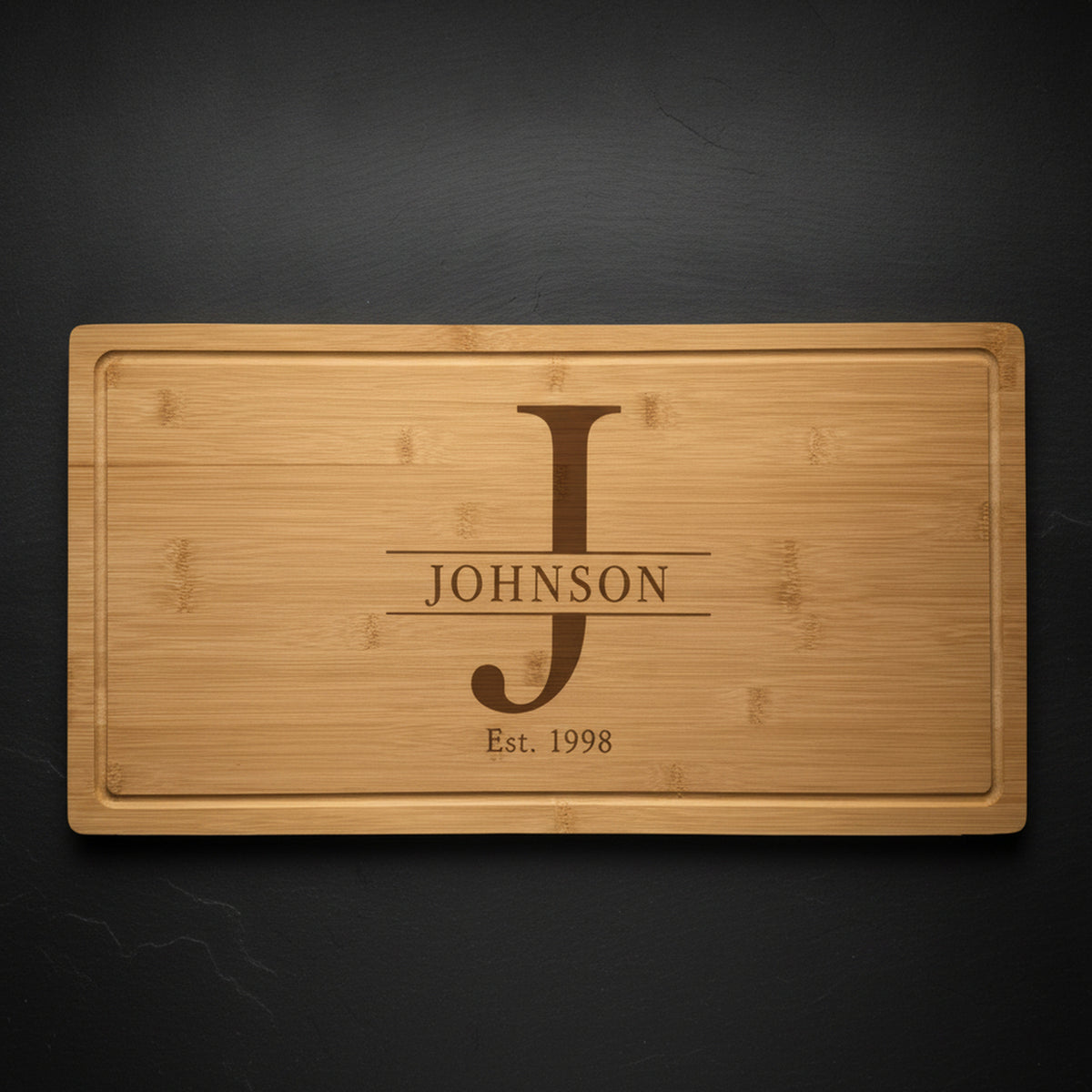 Personalized monogram cutting board in solid bamboo on dark slate, engraved with a tall serif J, the name JOHNSON across it and Est 1998 beneath.