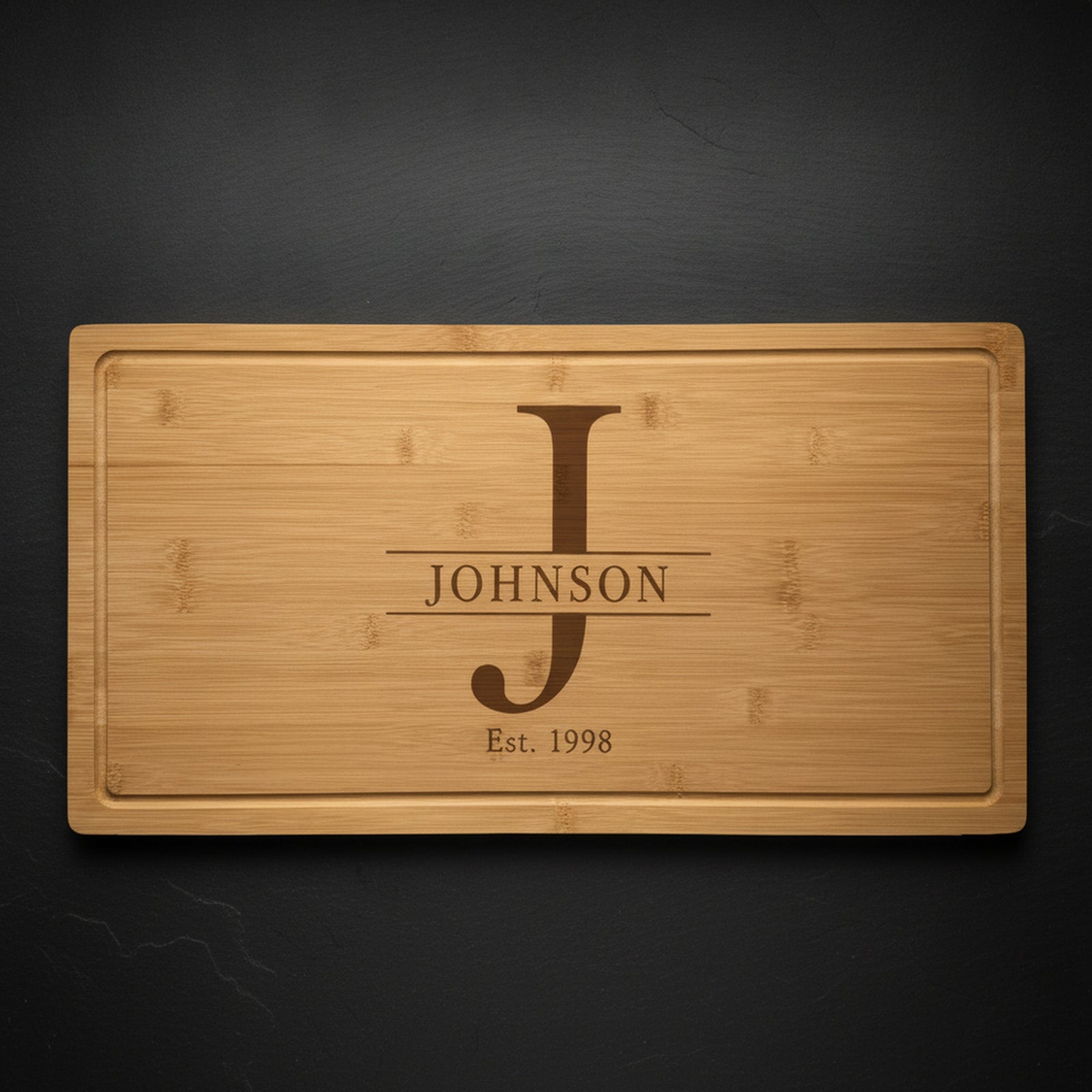 Personalized monogram cutting board in solid bamboo on dark slate, engraved with a tall serif J, the name JOHNSON across it and Est 1998 beneath.