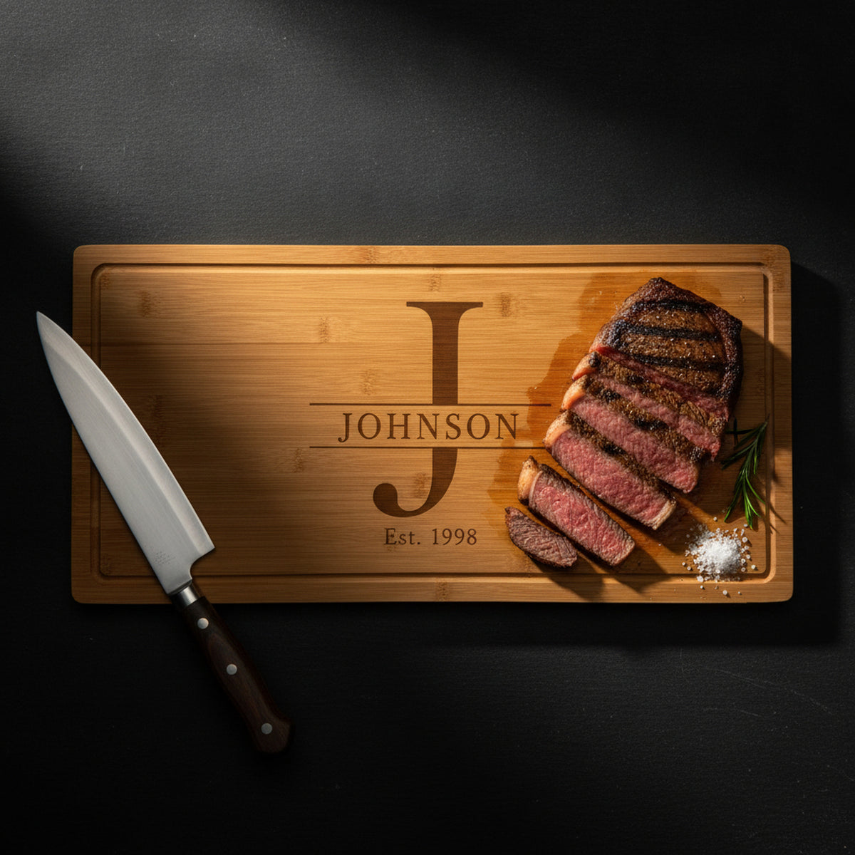 Large engraved bamboo carving board on dark slate holding a sliced medium rare ribeye with rosemary and salt, carving knife alongside, split monogram visible.