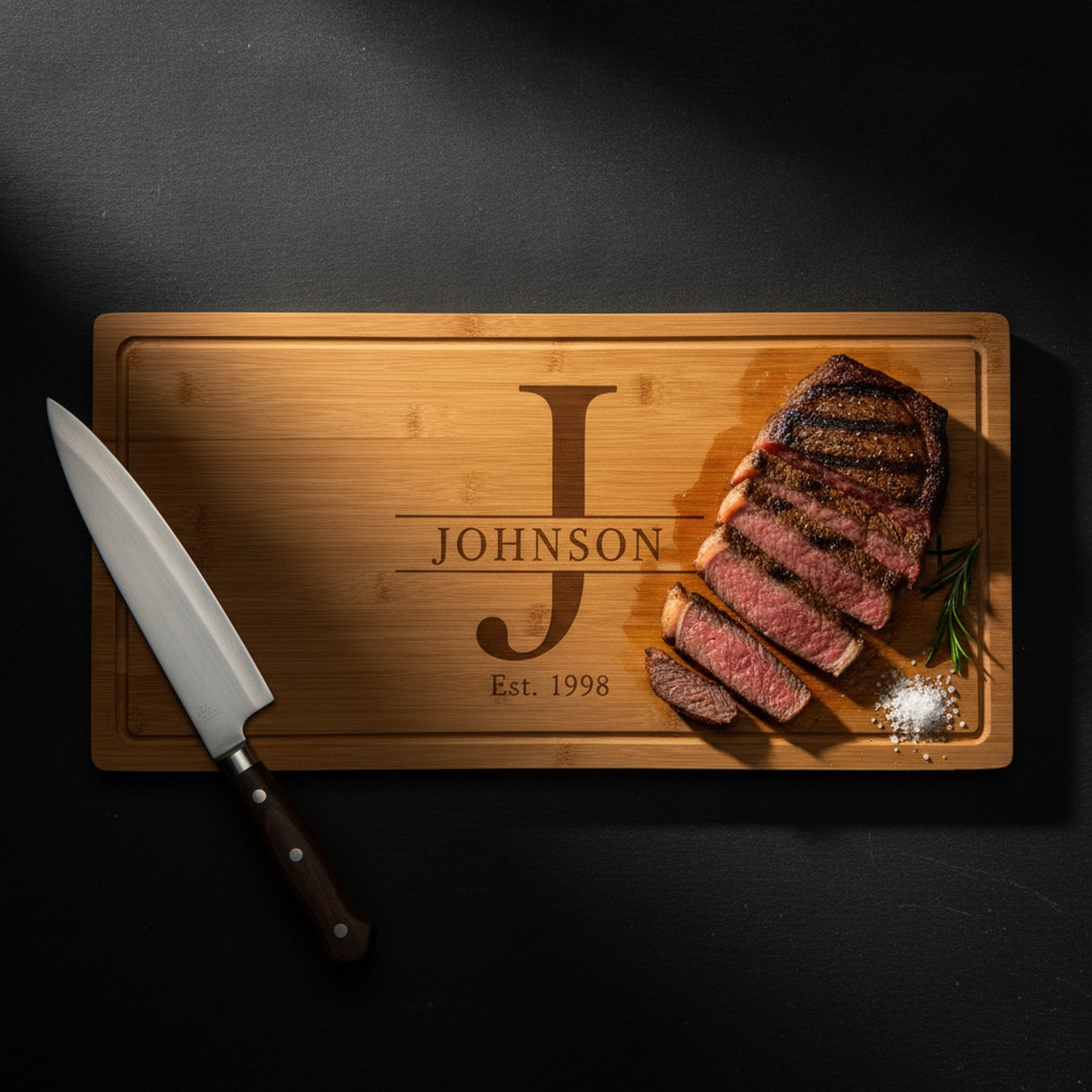Large engraved bamboo carving board on dark slate holding a sliced medium rare ribeye with rosemary and salt, carving knife alongside, split monogram visible.