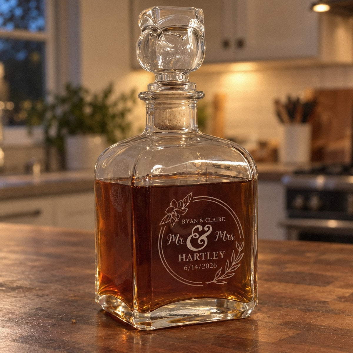 Personalized Mr and Mrs whiskey decanter etched RYAN & CLAIRE, HARTLEY and 6/14/2026 in a floral ring, filled with whiskey on a kitchen island