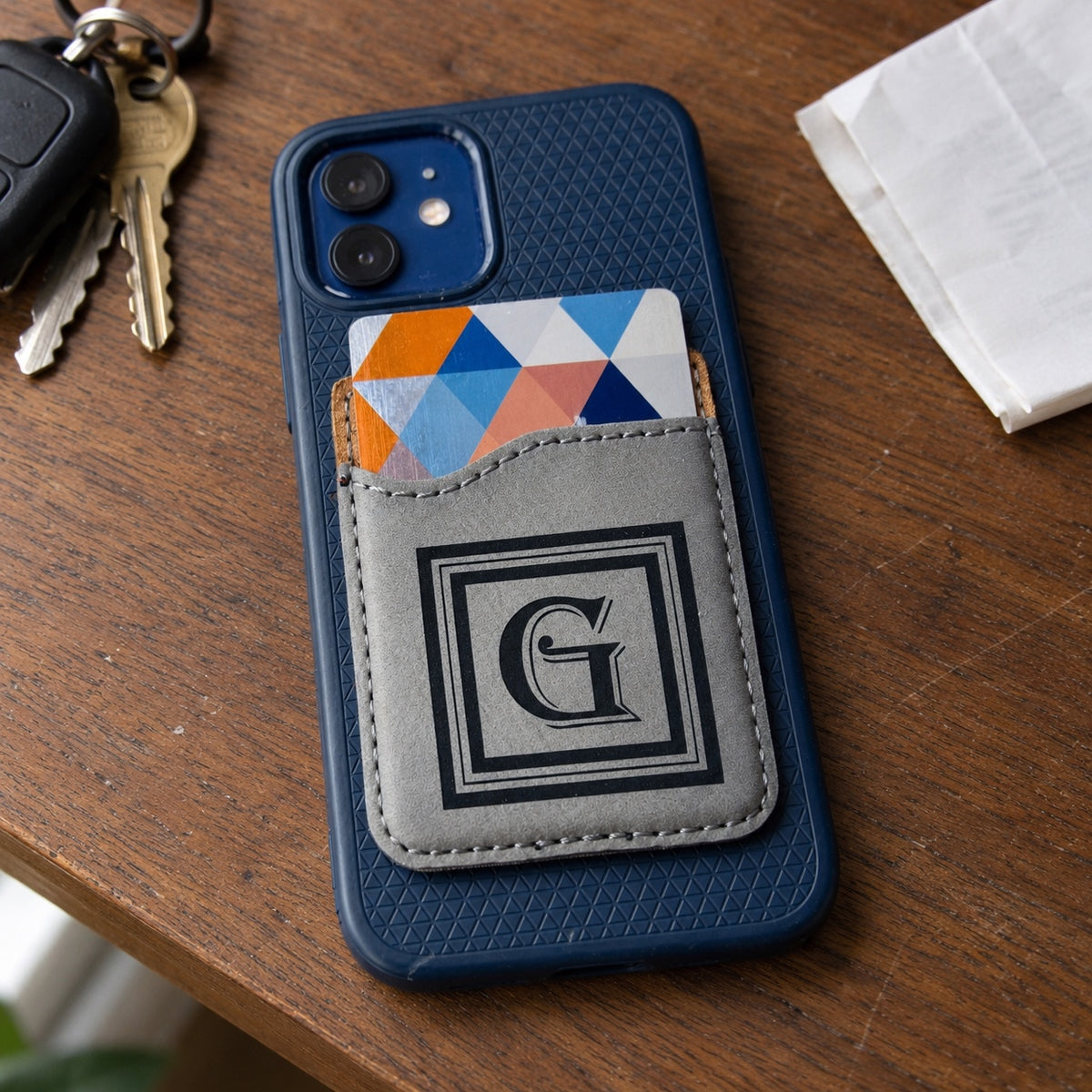 Personalized phone wallet in gray leatherette engraved with a square G monogram, stuck to the back of a navy phone on a walnut desk