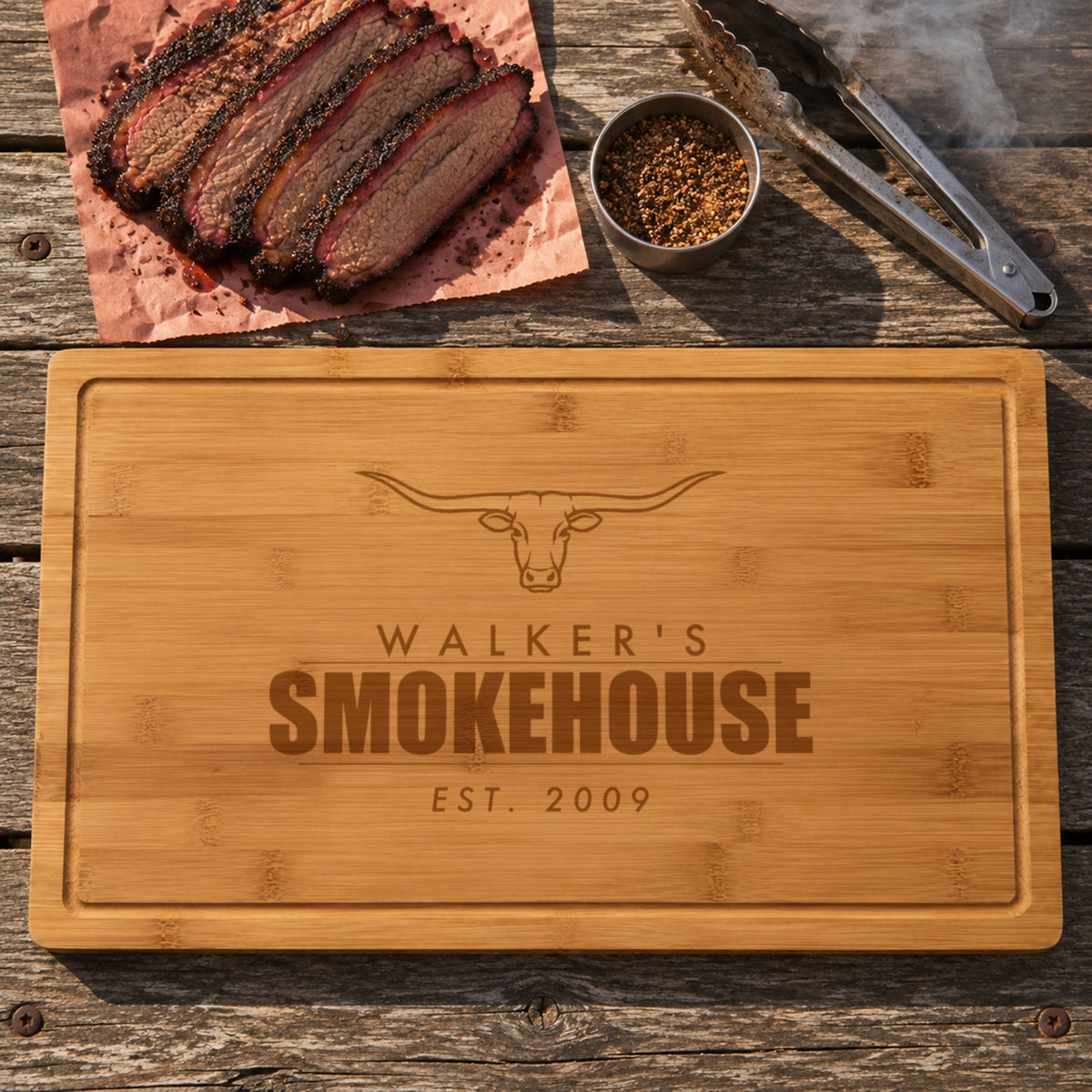 Personalized smokehouse cutting board engraved WALKER'S SMOKEHOUSE EST. 2009 under a longhorn, on a weathered picnic table beside sliced brisket, tongs and dry rub