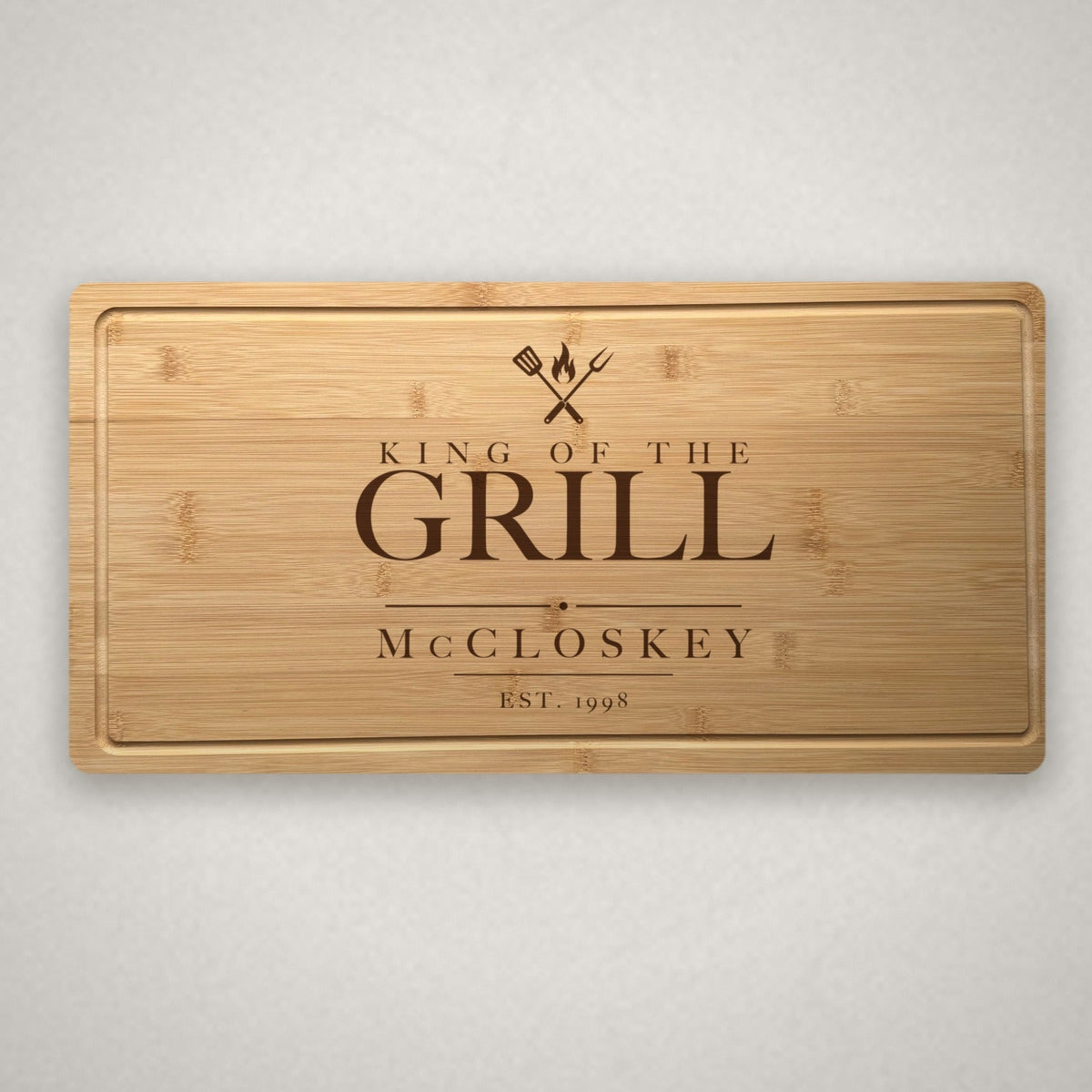 Personalized engraved steak board in solid bamboo on a light warm stone surface, engraved King of the Grill over a spatula, fork and flame with the family name.