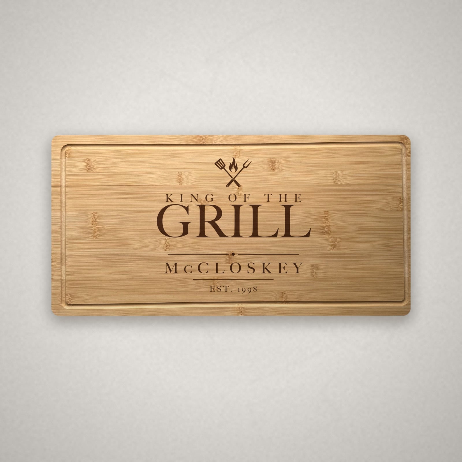 Personalized engraved steak board in solid bamboo on a light warm stone surface, engraved King of the Grill over a spatula, fork and flame with the family name.