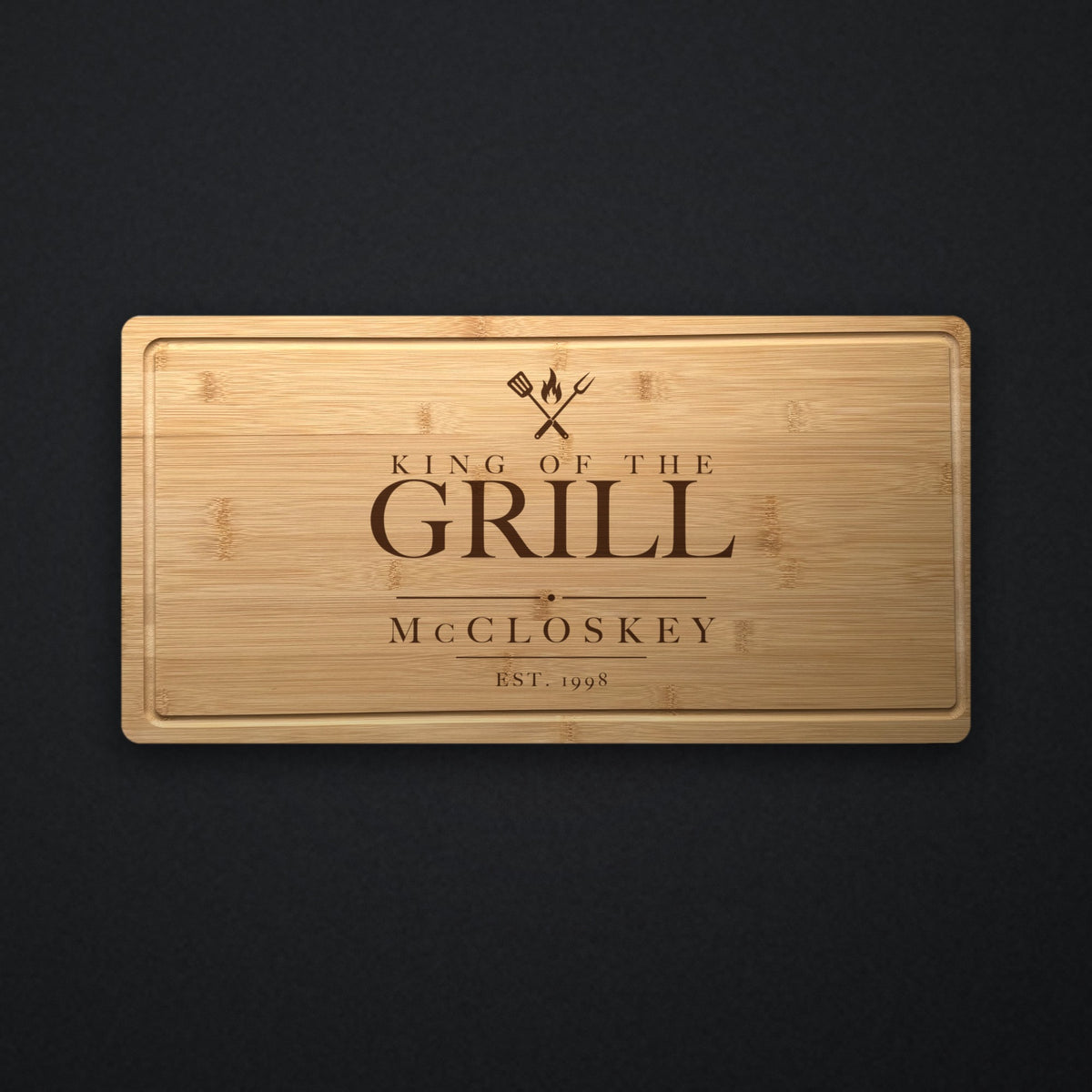 Personalized engraved steak board in solid bamboo on dark slate, engraved King of the Grill over a spatula, fork and flame with the family name beneath.