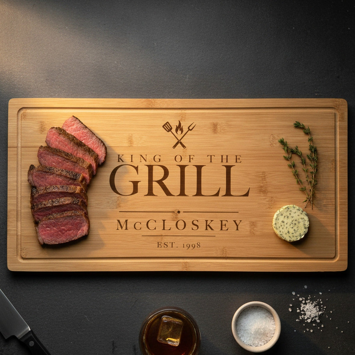 Engraved King of the Grill steak board on a dark slate counter with a sliced medium rare ribeye at one end, herb butter and thyme at the other, beside a steak knife.