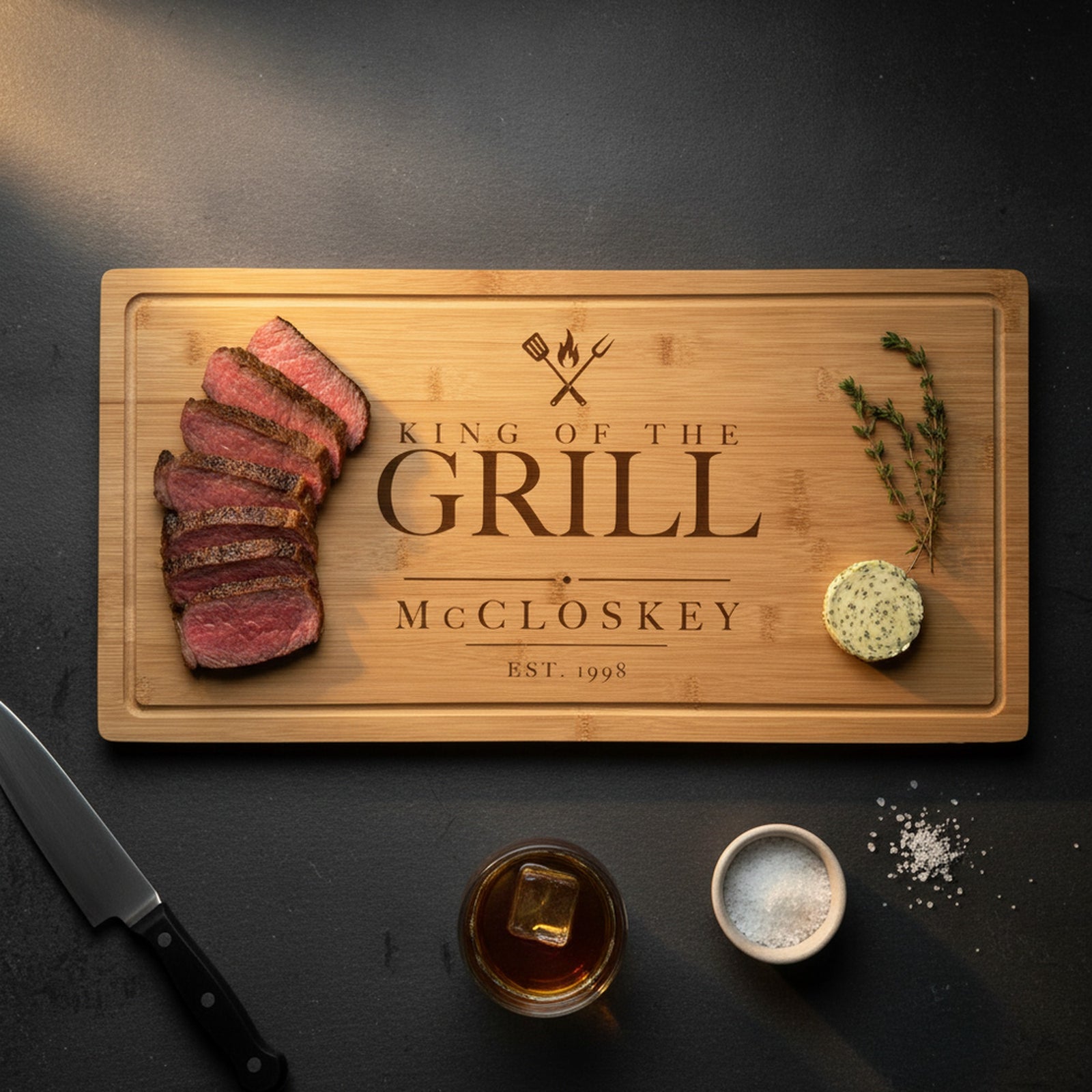 Engraved King of the Grill steak board on a dark slate counter with a sliced medium rare ribeye at one end, herb butter and thyme at the other, beside a steak knife.
