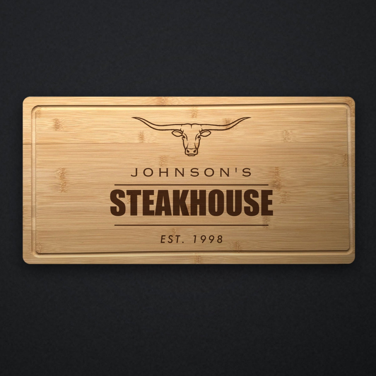 Personalized steakhouse cutting board in solid bamboo on dark slate, engraved with a longhorn steer above the name and the word Steakhouse.