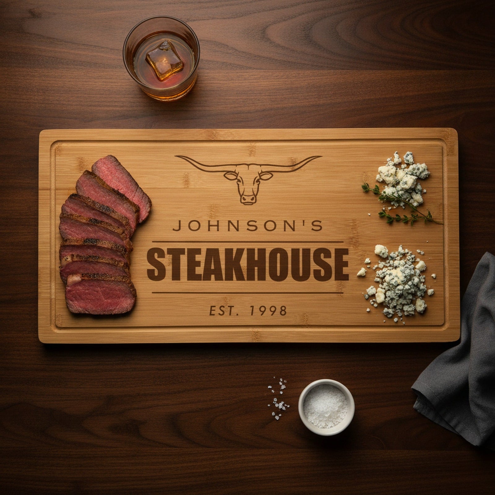 Engraved steakhouse bamboo board on a dark walnut bar top with sliced medium rare ribeye at one end, blue cheese and thyme at the other, beside a bourbon glass.