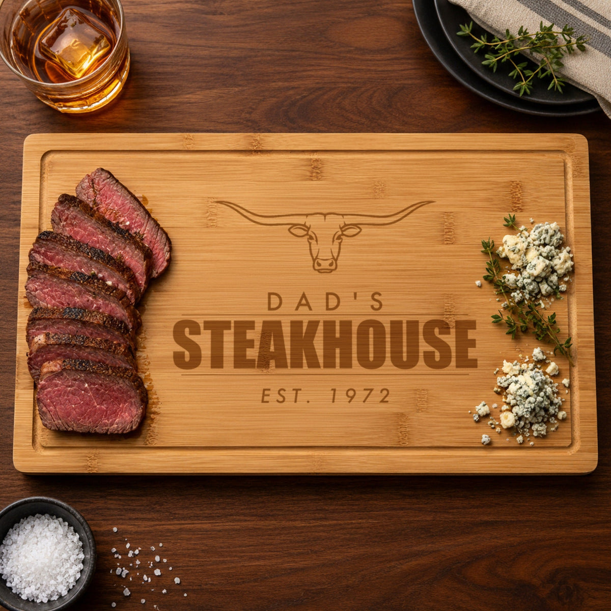 Personalized bamboo cutting board for dad engraved DAD'S STEAKHOUSE EST. 1972 under a longhorn steer, with sliced steak and blue cheese at the ends