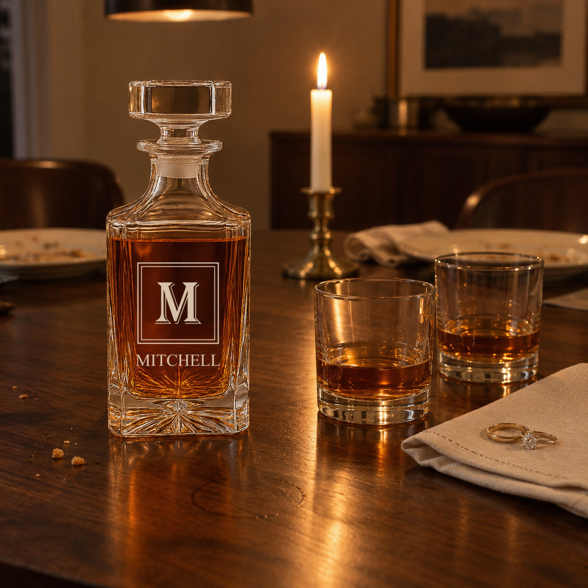 Personalized whiskey decanter engraved with an M monogram and MITCHELL beside two rocks glasses and her wedding rings after an anniversary dinner at home