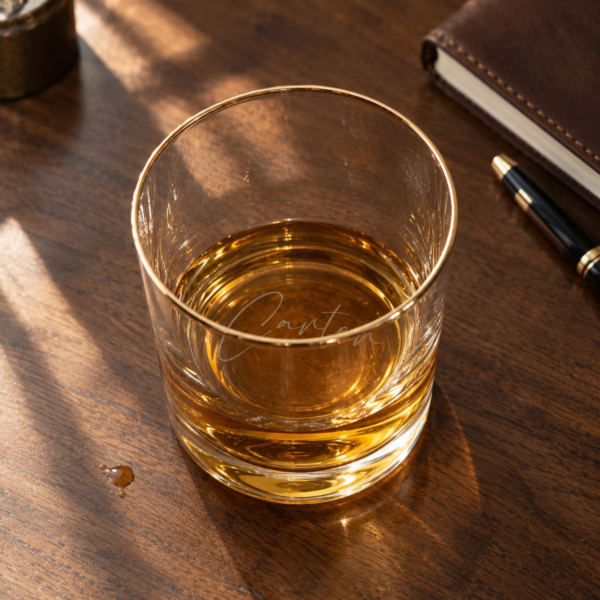 Personalized whiskey glass with a gold rim on a walnut desk, the name Carter engraved in script on the bottom and seen through a short pour of bourbon