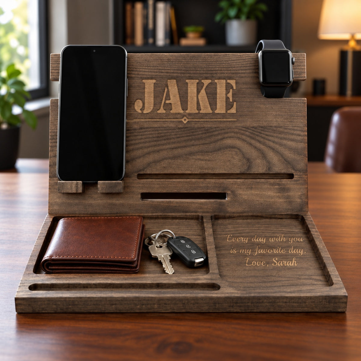 Wooden charging dock with JAKE laser engraved on the back panel and a love note signed Sarah in the tray, holding a phone, smartwatch, wallet and keys on a desk