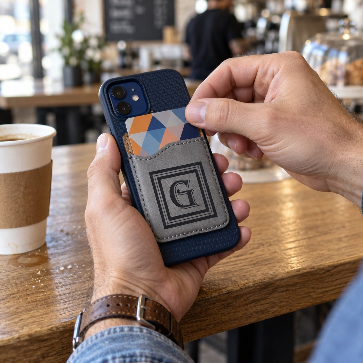 A man slides a card out of his monogrammed phone wallet at a coffee shop counter, a slim personalized gift for him