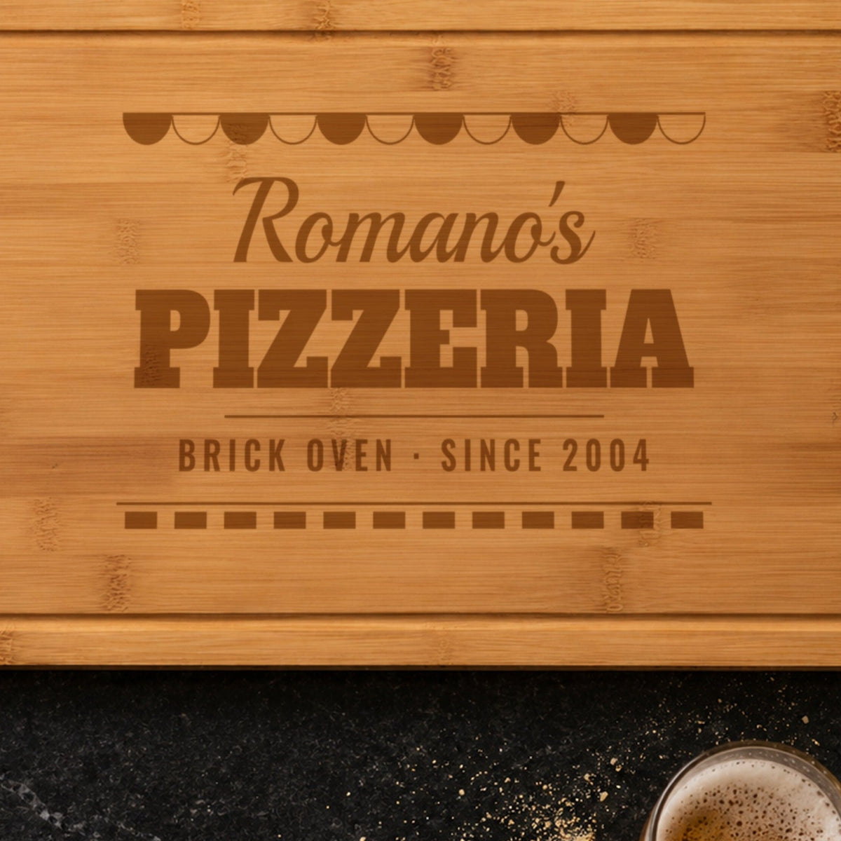 Close-up of the laser engraving on the pizza cutting board: the striped awning, Romano's in script and PIZZERIA burned into the bamboo grain