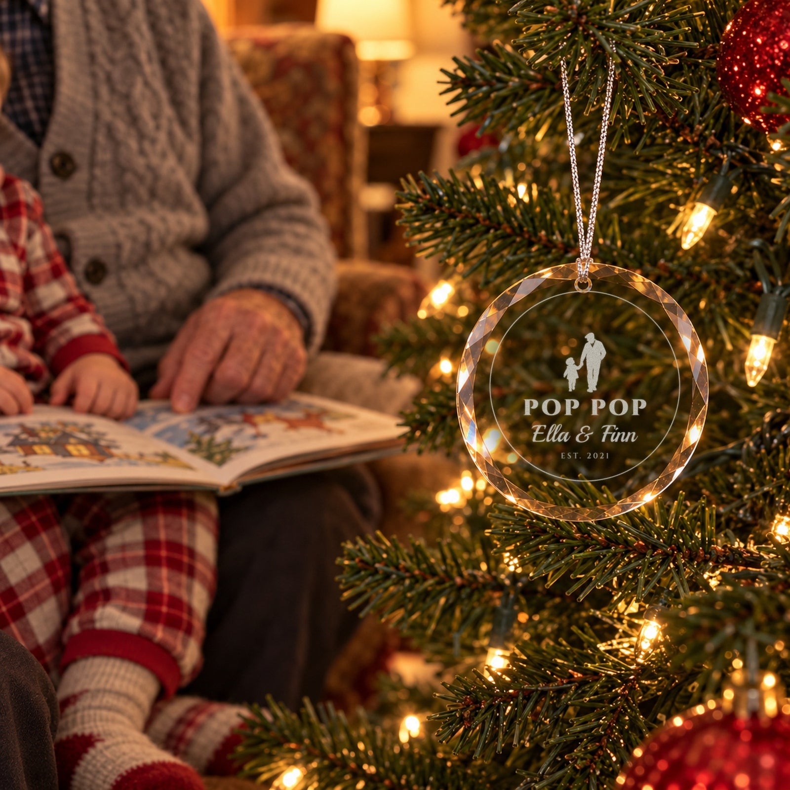 Grandfather in a cardigan reading a picture book to his grandchild by the Christmas tree beside the engraved glass ornament reading POP POP, Ella & Finn, EST. 2021