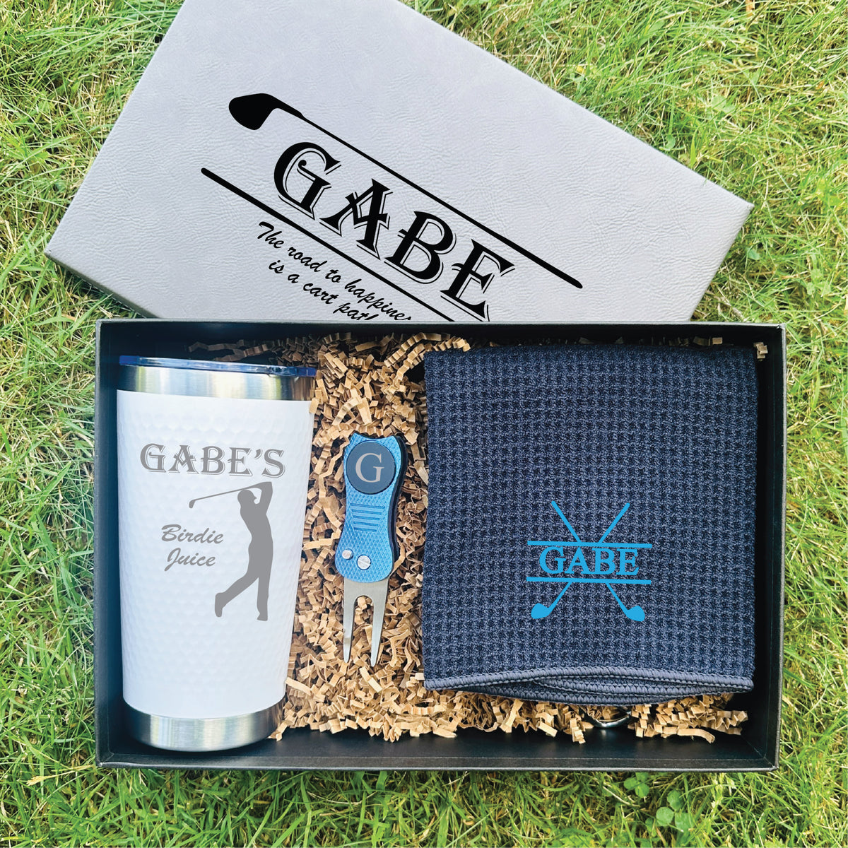 Personalized golf gift set in black presentation box containing stainless steel tumbler engraved with sample name "GABE'S Birdie Juice," embroidered navy towel with crossed clubs design, blue divot tool, and carabiner clip on kraft paper filler