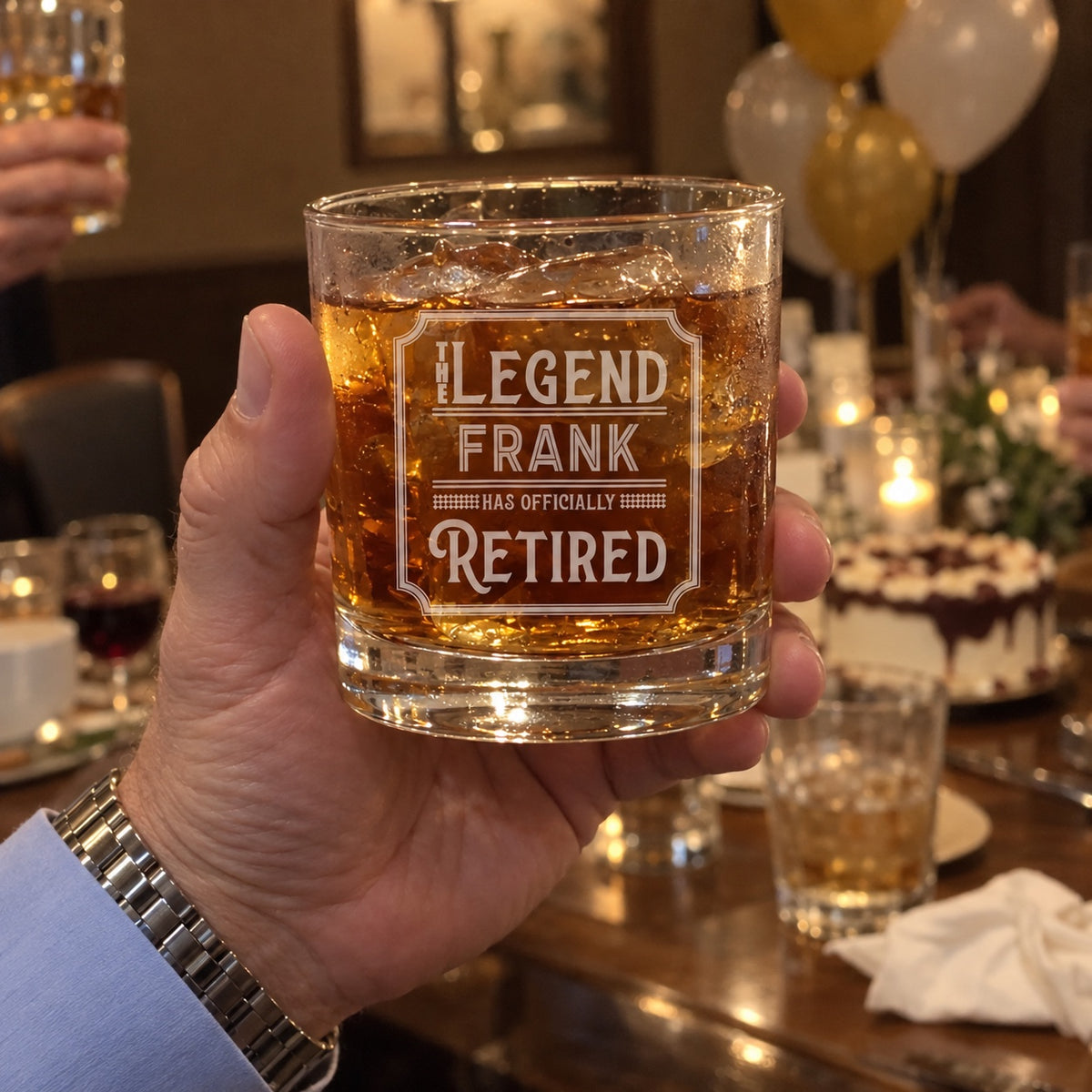 A retiree raises his engraved whiskey glass reading THE LEGEND FRANK HAS OFFICIALLY RETIRED in a toast at his retirement party, cake and balloons behind.