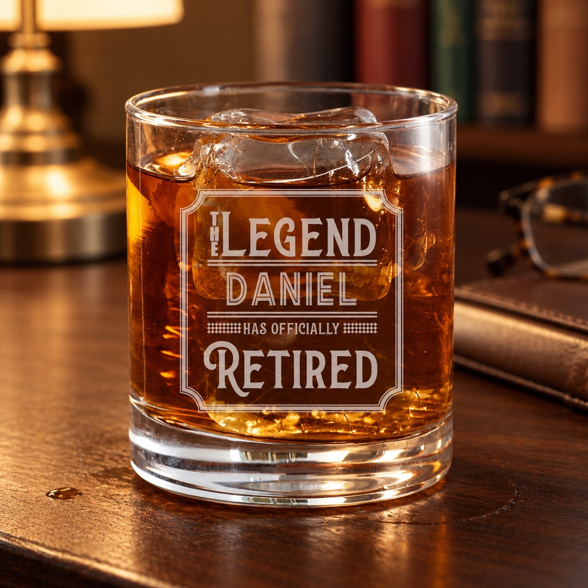 Retirement whiskey glass engraved THE LEGEND, DANIEL, HAS OFFICIALLY RETIRED in a vintage frame, a personalized rocks glass of whiskey on a study desk.